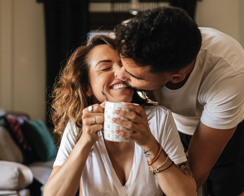 Woman holding a coffee mug while her partner gives her a kiss